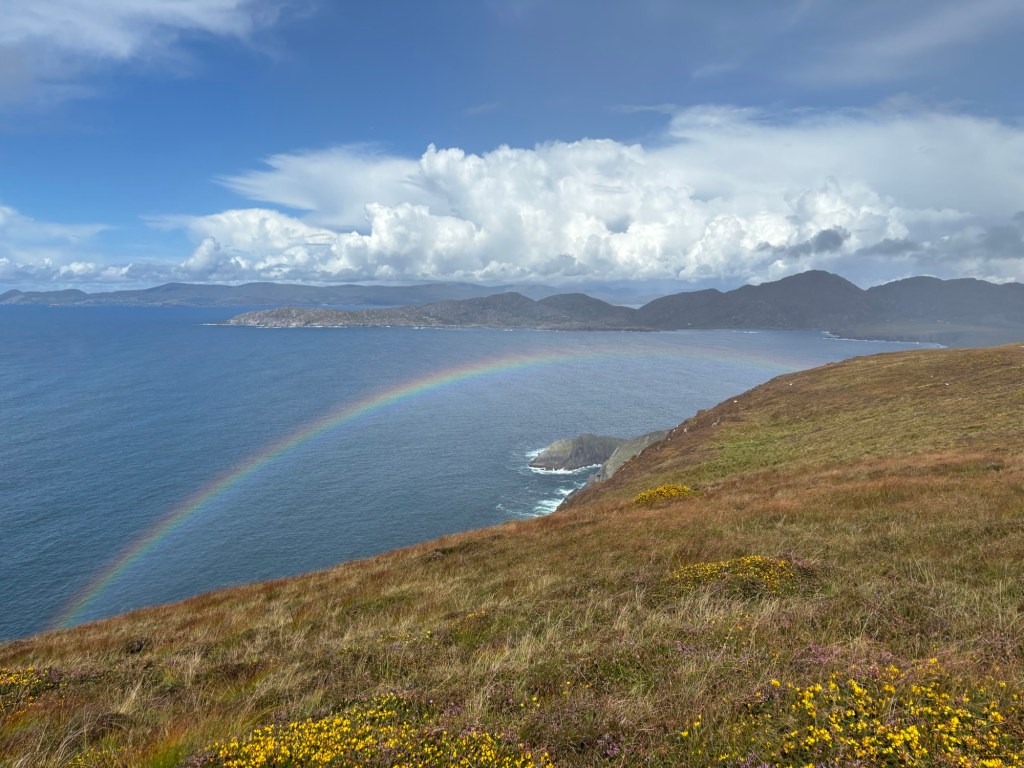 Photo off the coast of Ireland showing a hill with clouds and a rainbow above a blue bay.
